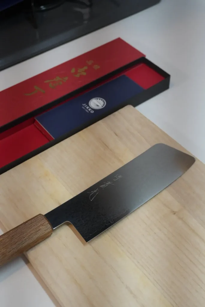 JIKKO Damascus nakiri knife showing the flat rectangular blade profile on a hinoki cutting board